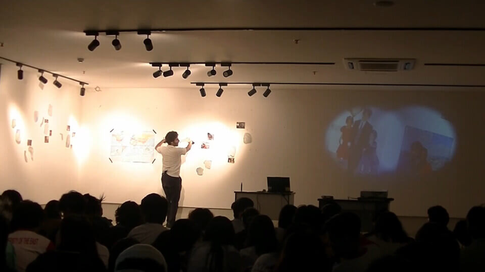 A crowd sits in a dark gallery space. Ceiling lights illuminate the left part of the wall, which is adorned with photos and map fragments. The crowd watches as the performer (Stephan E. Perez) points his arms, with cameras attached, towards the photos and maps. He looks towards the right side of the wall, where the two cameras' feed is being projected in the shape of two overlapping circles, blurred at the edges. The left circle shows a young boy and two even younger girls. The right circle shows two adults in front of a landscape.