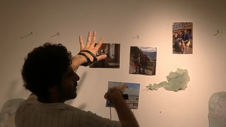 The performer points his hands, with cameras taped inside of the palms, towards the wall. The wall has photos on it taken in Austria and Poland and a map of Austria
