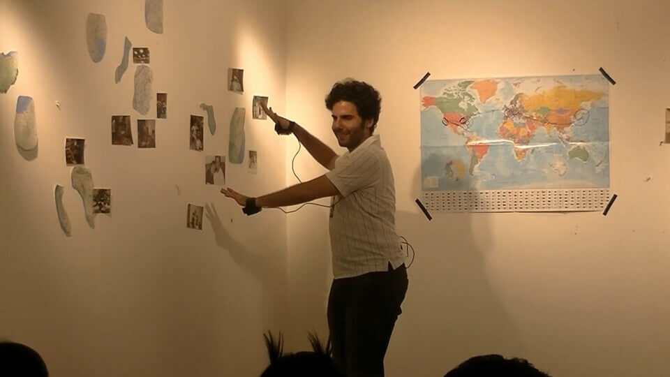The performer smiles and points his hands, with cameras taped inside of the palms, towards the wall. The wall has photos on it and map fragments of various U.S. states and cities. The perpendicular wall behind the performer has a complete world map with some lines and circles drawn in marker.