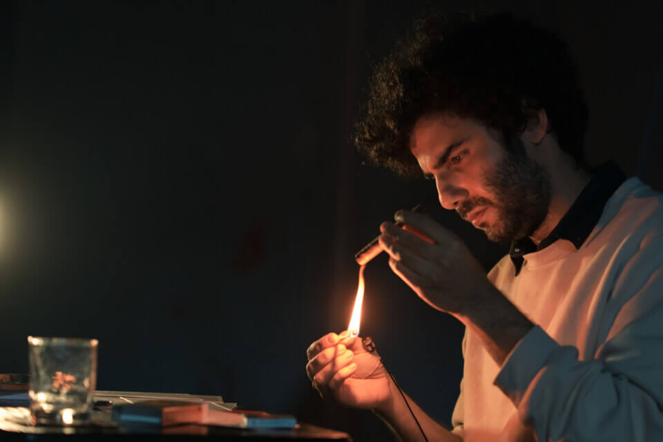 The performer, stephan e perez, sits at a small folding desk and lights a cigar with a match. The light from the match illuminates his face and upper body. A microphone is taped to his wrist