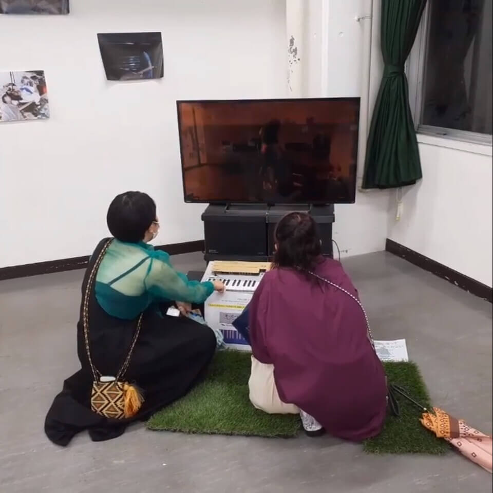 Two women crouch on squares of artificial grass and press keys of a MIDI piano. Behind the piano is a TV monitor showing composited images of performances from events organized by the tokyo performance art platform, ham tamago sandwich