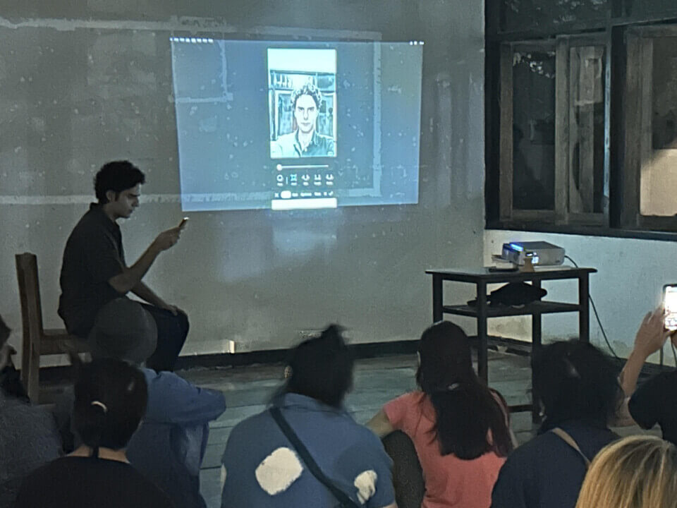 The performer sits on a chair using his phone. The photo of him that he is editing is being projected. The audience watches.