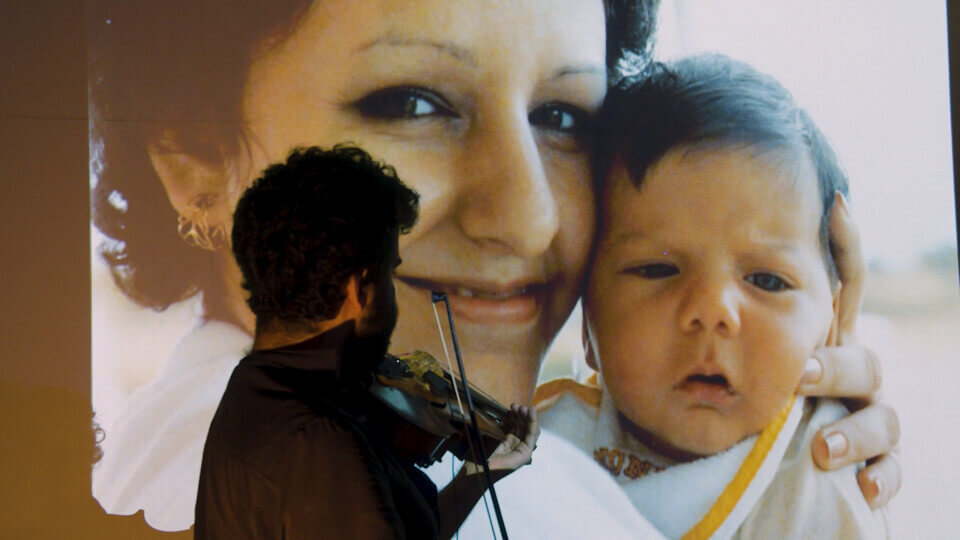 the performer plays viola in front of a projection of a woman in her 30s holding a baby