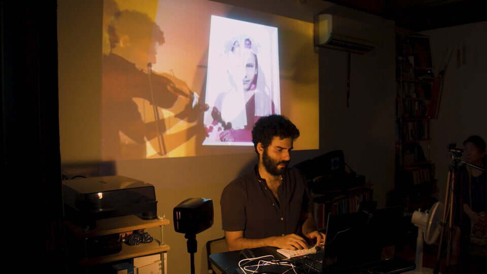 the performer sits in front of a laptop and types. Behind him, there is a wall projection of an alpha-composition of footage of his viola-playing. This footage also shows the wall projections of photos including one of a young girl less than 10 years old and a wedding photo