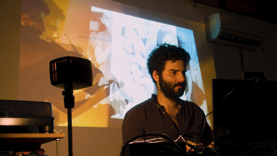 the performer sits in front of a laptop and types. Behind him, there is a wall projection of an alpha-composition of footage of his viola-playing. This footage also shows the wall projections of some old, monochrome family photos
