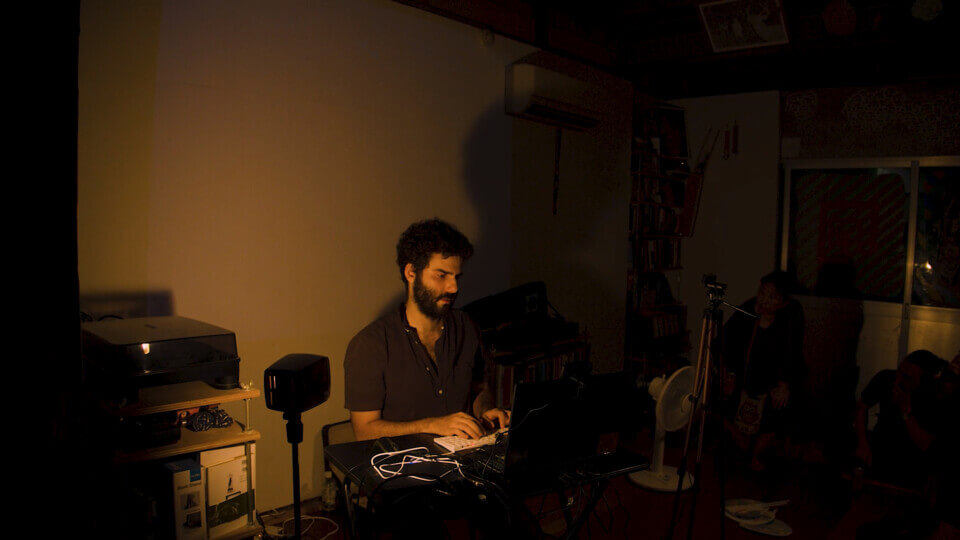 the performer sits in front of a laptop and types. Behind him, the wall is blank