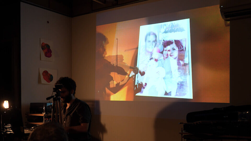 the performer sits in front of a laptop and types. Behind him, there is a wall projection of an alpha-composition of footage of his viola-playing. This footage also shows the wall projections of some old, partly monochrome family photos