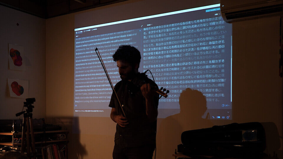 the performer fingers a MIDI violin in preparation to play. Behind him, there is a wall projection with an additive alpha composition of multiple images of screen-captured text in English and Japanese