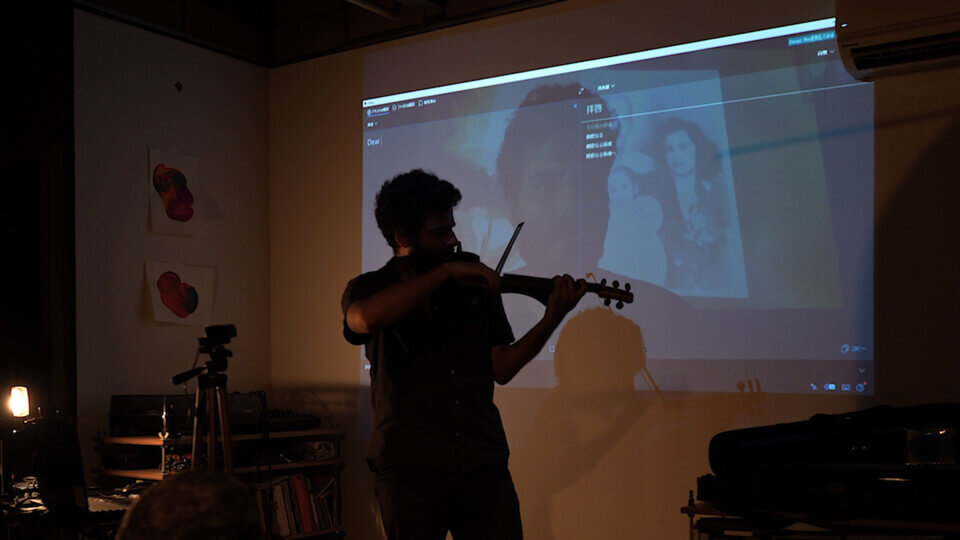 the performer plays a MIDI violin in front of a wall projection. The projection shows an additive alpha composition consisting of several images. Recognizable elements are English and Japanese text from a screen capture, the performer's closeup, and a monochrome photo of a young couple with a baby