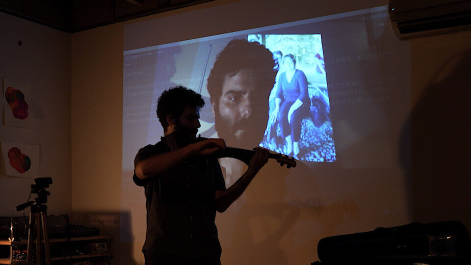 the performer plays a MIDI violin in front of a wall projection. The projection shows an additive alpha composition consisting of several images. Recognizable elements are English and Japanese text from a screen capture, the performer's closeup, the performer playing viola and a photo of a teenage boy with a woman in her 50s