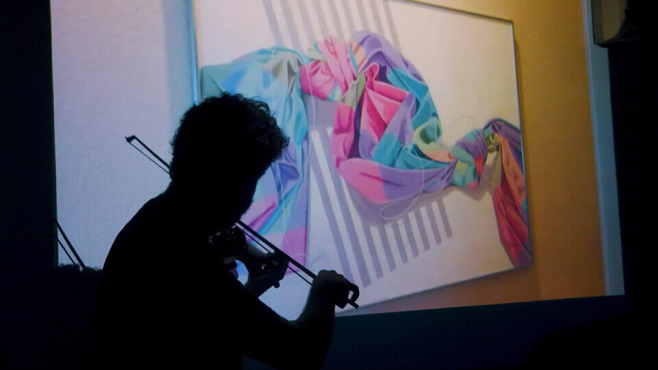 sillhouette of the performer, stephan e perez, playing MIDI violin in front of a projection. The projection shows a painting of a colorful, abstract cloth tied into knots