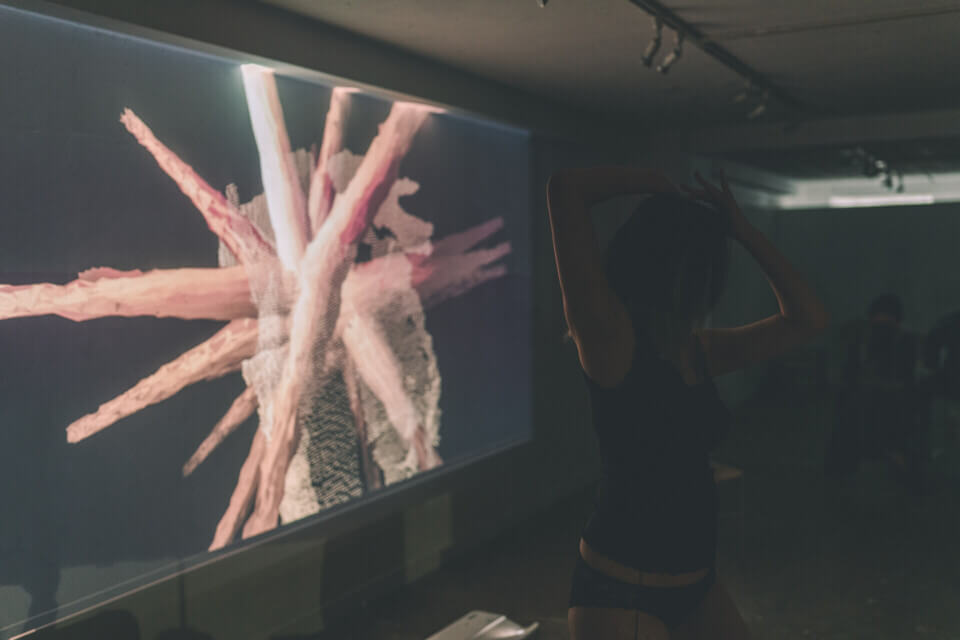 a woman in black underwear and a black top, the performer cat-o-nine-tails, holds her hands above her head. Behind her, a wall projection shows a 3d cloud-point representation of her body superimposed with a floating, 3d spiky form. The spikes are long and have a ripply texture