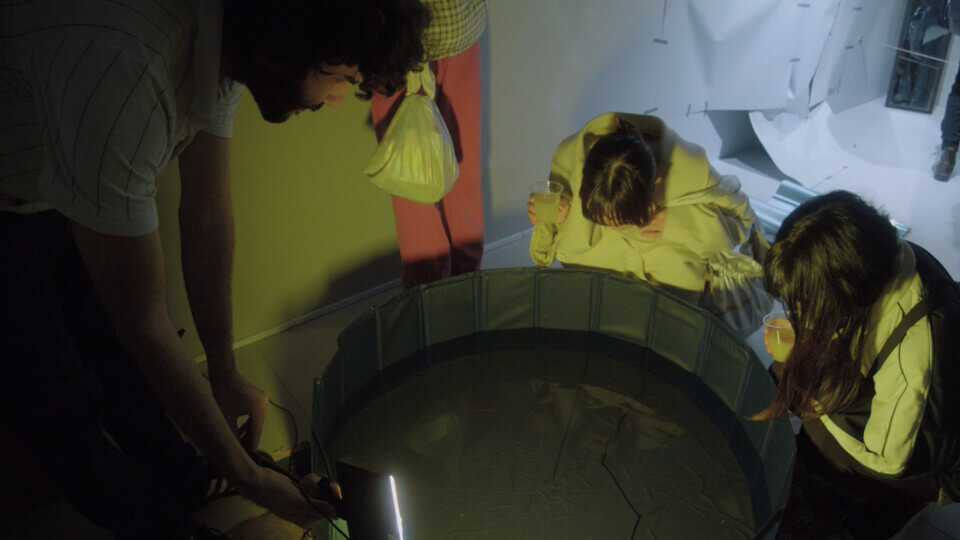 The performer, stephan e perez, leans over a small, dimly lit, vinyl pool filled with water. Two women are crouching looking into the water. Reflections of people are visible in it.