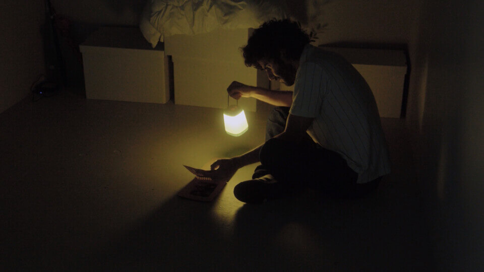 In a dark room, the performer sits cross-legged and holds a lantern in one hand, turning through a photo album with another