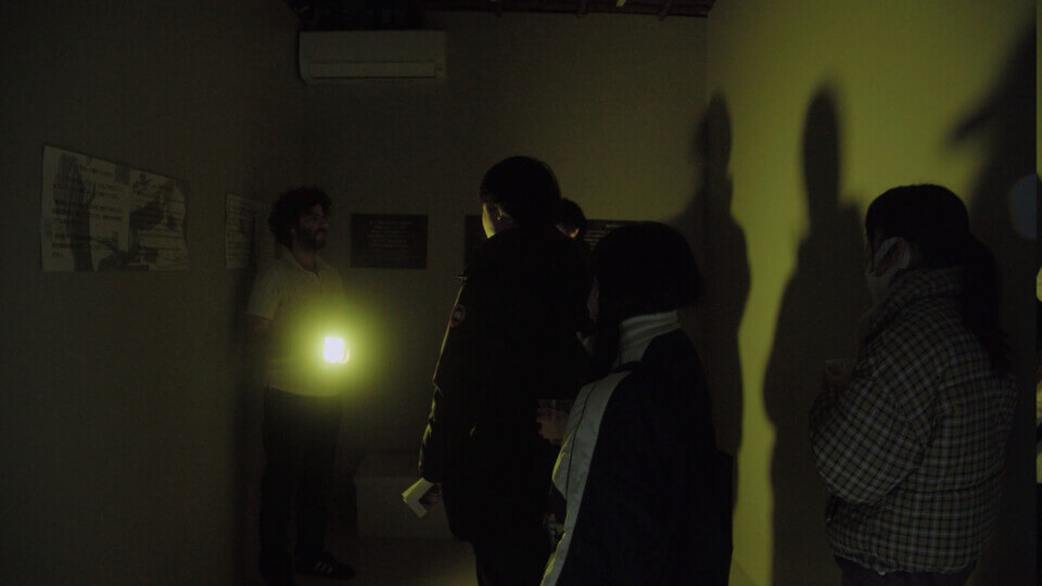 the performer aims the yellow light of a handheld lamp at event guests, casting their shadows on the wall