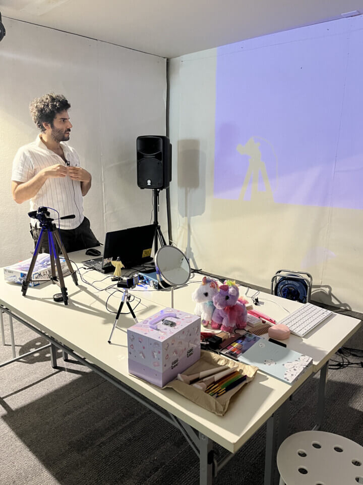 stephan e perez looks at the projection in preparation for the event. In front of him is a large desk. On it are: the unicorn makeup kit, makeup brushes, notebooks, combs, two unicorn plushies, a mirror, a laptop, two cameras on tripods and a gunpla box