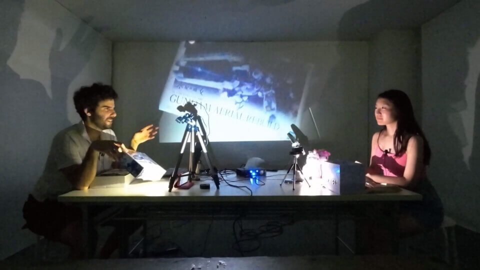 The performers, stephan e perez and akari, are seated at both ends of the long table. One camera is pointed towards Akari, the other is pointed downwards towards the table space in front of Stephan. On the wall behind the, a projection shows the gunpla box which Stephan is tilting up towards the camera
