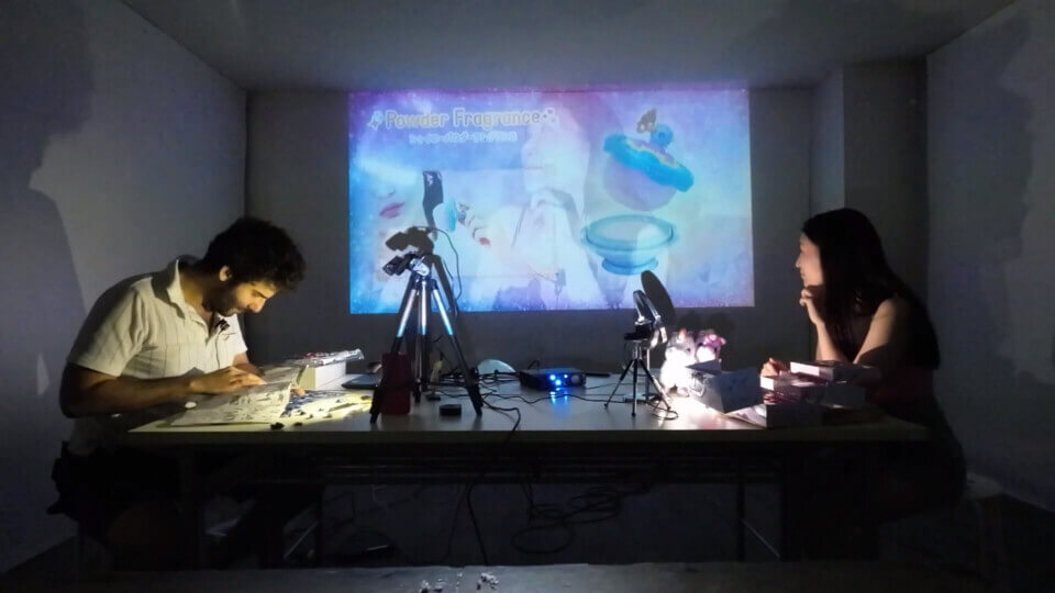 stephan consults the gunpla instructions while akari looks at the projection, which shows a composite of her and an advertisement for a makeup kit for little girls