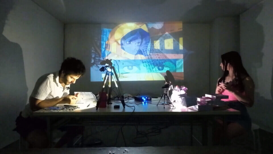 stephan works on the gunpla and akari looks at the projection, which shows a composite of stephan's work on the gunpla and a scene from the anime Mobile Suit Gundam