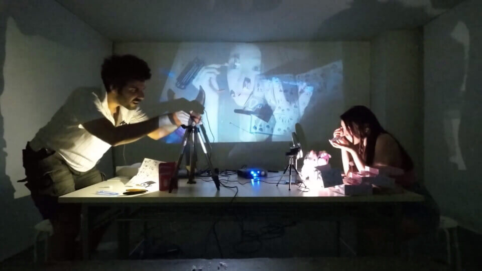 stephan adjust his camera while akari applies makeup. The projection shows a composite of akari and the gunpla workspace