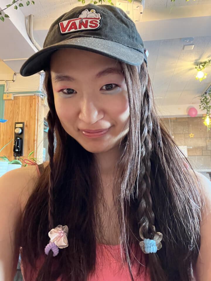 akari, smiles at the camera after the performance. Her face is made up with kids' makeup- glossy pink lips and blue & pink eyeshadow. Part of long dark hair is braided and she sports a baseball cap.
