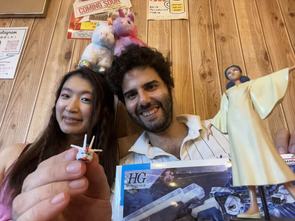 akari and stephan, smiling, each balance a unicorn plushy on their head, with the unicorns leaning against each other. In front of them are the gunpla box and a figurine of Lalah Sune, a character from the original Gundam series and movies. Stephan's hand is in front, delicately holding the completely head of the gundam.