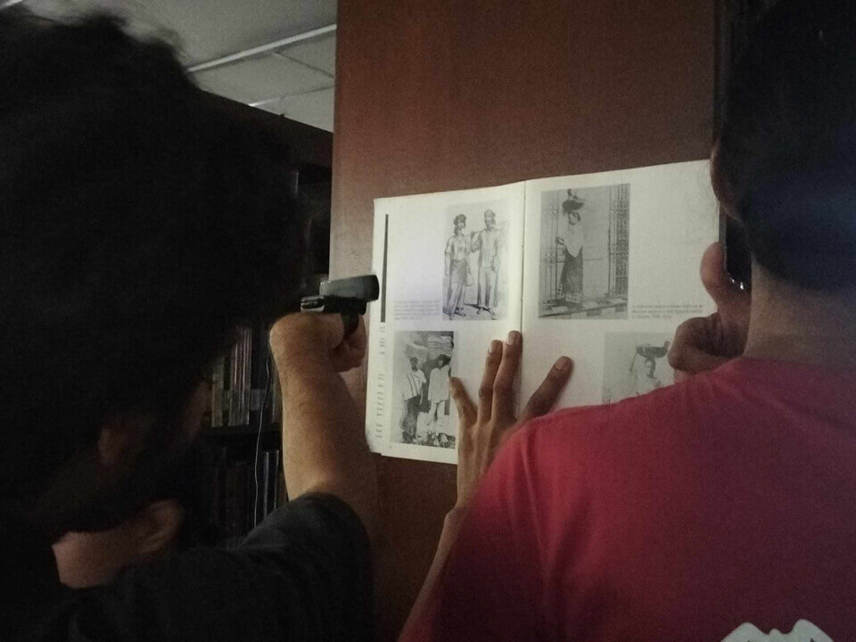 The performer points his fist, which has a webcam taped to the top of it, at a book, held open by a man against the side of the bookshelf. The book has old pictures of Philippino farmers and workers in traditional attire.