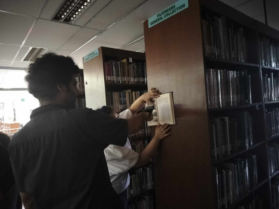 The performer points a camera at a book, held by someone against the side of a bookcase