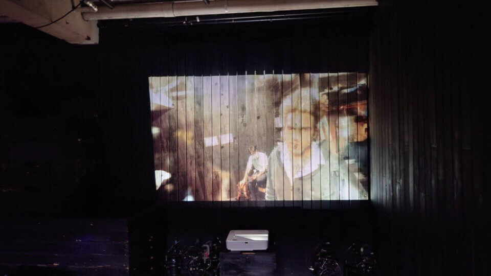 projection on the black, planked walls of Subterranean. The image shows previously recorded event guests, one of them looking towards the camera