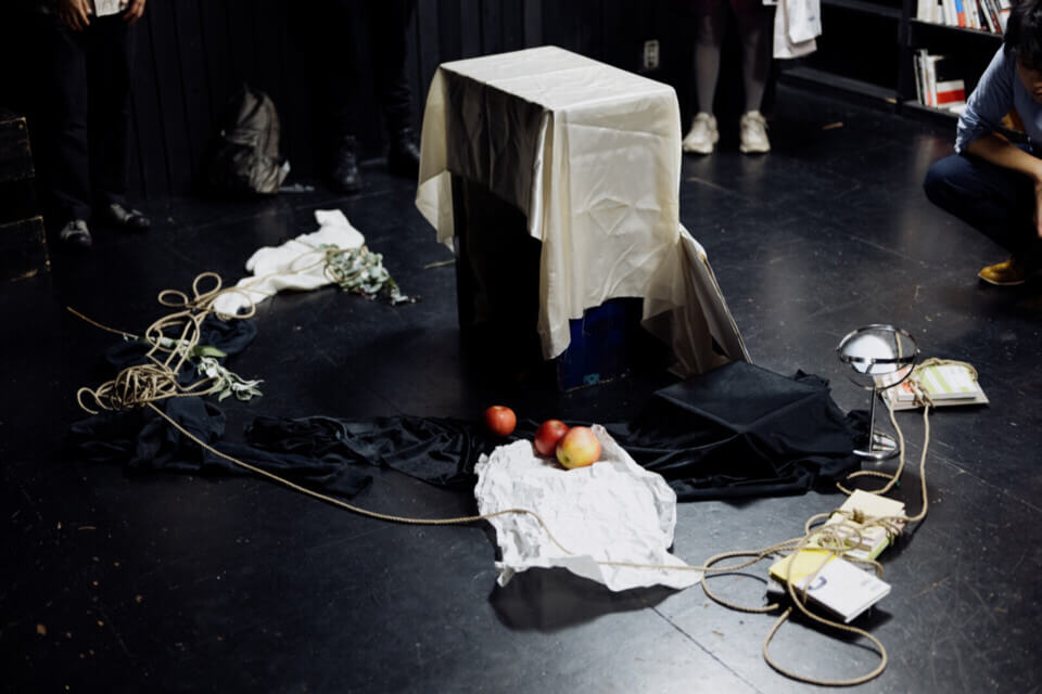 the table has the following items strewn about it: rope, apples, crumpled paper, books tied into the rope, flowers, a mirror. The feet of event guests are seen standing around these items.