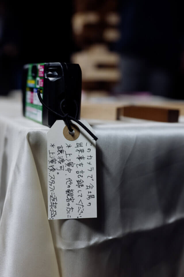 a disposable camera sits next to some stray jenga blocks on a table, the jenga tower in the background. The camera has a tag which urges the event guests, in Japanese, to take phots during the event