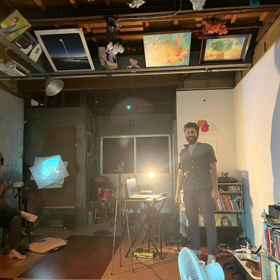 On the right, the performer stands by the laptop computer, smiling. At his feet is the viola case. A camera tripod is next to the computer. On the left, the canvases and an event guest can be seen